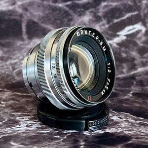 May include: A silver and black camera lens with the text "Jupiter-8M" and other markings. The lens has a ridged focus ring and aperture settings. The lens is resting on a black lens cap. The lens is a 5 cm f/2 lens.