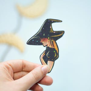 Peut inclure: Un autocollant de sorcière avec un chapeau et une robe noirs avec des accents de paillettes. La sorcière regarde vers le bas avec une expression triste.
