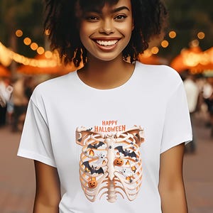 Può includere: T-shirt bianca con grafica a tema Halloween. Il design presenta una gabbia toracica con pipistrelli, zucche e fantasmi. Le parole "Happy Halloween" sono stampate sopra la gabbia toracica. La maglietta è a maniche corte.