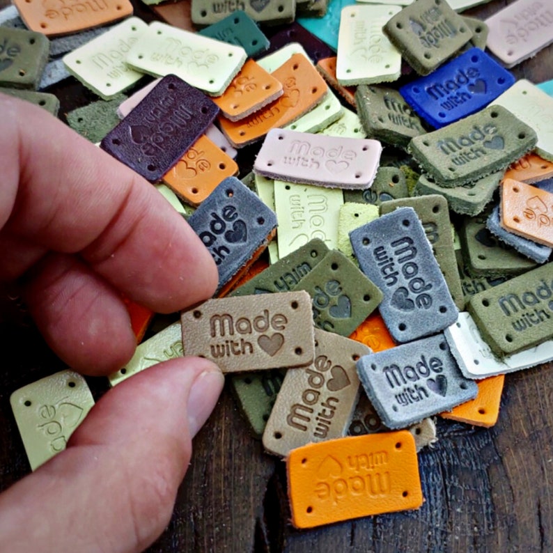 May include: A close-up of a variety of leather labels with the text "Made with love" printed on them. The labels are in different colors, including brown, green, blue, and gray. Some of the labels have a heart shape.