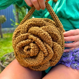 May include: A handcrafted, round, crochet bag in a warm, golden-brown hue. The bag is designed to resemble a rose, with spiraling layers creating a floral pattern. It features a braided handle for carrying.