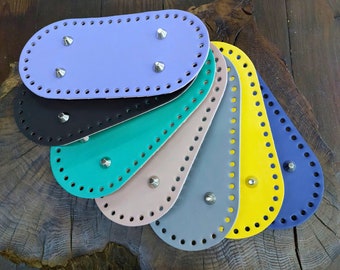Op de afbeelding: Een stapel van zeven ovale lederen stukken in verschillende kleuren, waaronder paars, zwart, teal, beige, grijs, geel en blauw. Elk stuk heeft een rij gelijkmatig verdeelde gaten rond de rand en metalen klinknagels in het midden.