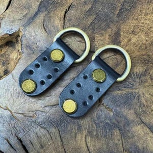 Könnte beinhalten: Zwei schwarze Lederriemen mit Messingringen und Messingnieten. Die Riemen haben ein gepunktetes Muster.