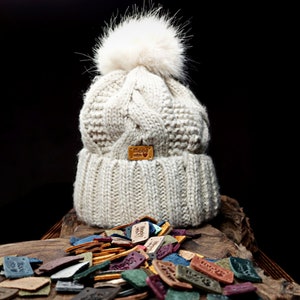May include: A white knitted beanie with a fluffy pom pom and a brown leather label that says "Made with love". The beanie is surrounded by a variety of colorful leather labels.