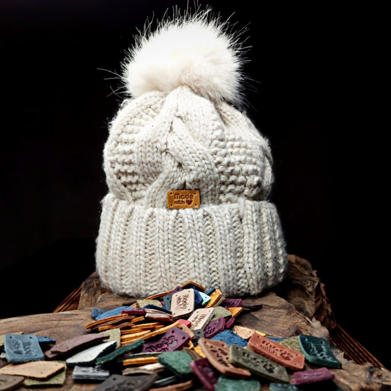 May include: A white knitted beanie with a fluffy pom pom and a brown leather label that says "Made with love". The beanie is surrounded by a variety of colorful leather labels.