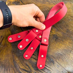 May include: A red leather harness with multiple straps and silver-tone hardware. The harness is held by a hand, showcasing its design and construction. The leather appears to be smooth and the overall aesthetic is modern.