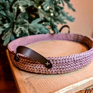 Könnte beinhalten: Ein runder, gehäkelter Korb in Staubrosa mit Holzbasis. Der Korb hat zwei dunkelbraune Ledergriffe mit einem weißen Kreuzstichdetail. Der Korb steht auf einer Holzoberfläche.