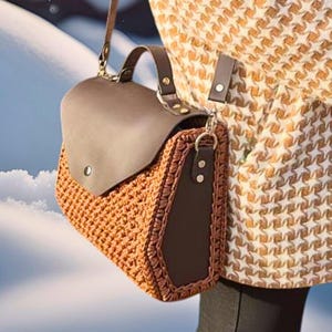 May include: A brown crocheted bag with a brown leather flap and a brown leather strap. The bag is being worn by a person wearing a brown and white patterned sweater.