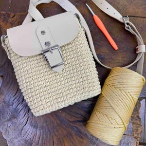 Peut inclure: Un sac bandoulière crocheté de couleur crème avec un rabat en cuir lisse, une boucle en métal argenté et une bandoulière réglable. À côté du sac se trouvent une bobine de cordon crème assorti et un crochet à manche orange pour le travail.