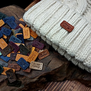 May include: A close-up of a white knitted hat with a brown leather tag that reads "Made with Love". The hat is surrounded by a variety of colorful leather tags with different designs and text, including "Made with Love" and "Yarn Love".