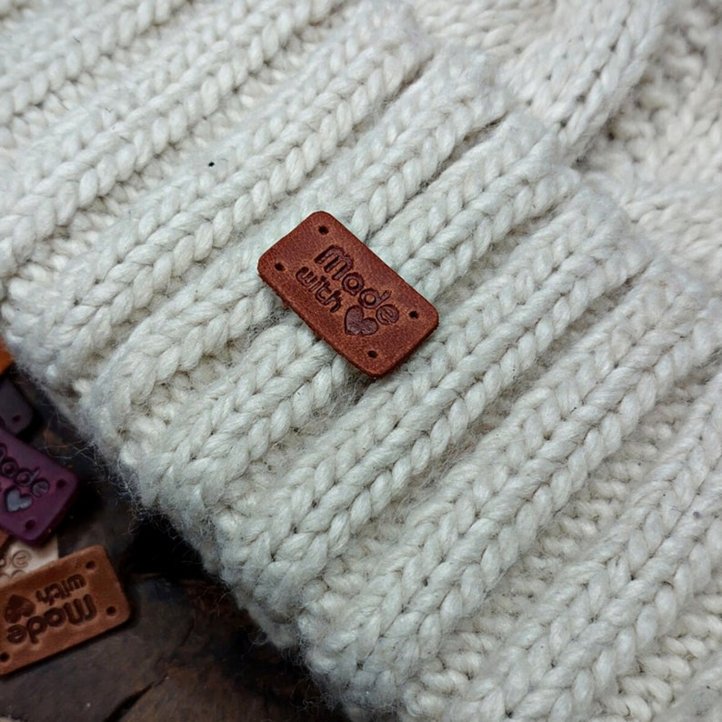 May include: A close-up of a white knitted beanie with a brown leather patch that says "made with" and a small heart.