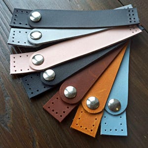 May include: A set of eight leather straps with metal buckles. The straps are in various colors, including black, brown, tan, light blue, and pink. The straps are all the same size and have a hole pattern on one end.