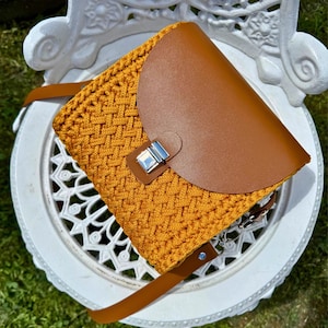 May include: Mustard yellow crochet crossbody bag with a brown leather flap and strap. The bag features a silver-colored clasp and is sitting on a white decorative chair.
