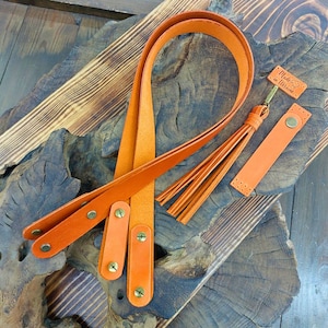 May include: A set of two orange leather straps with metal hardware and a tassel. The straps are on a wooden surface with a tag that says "Made in Ukraine".