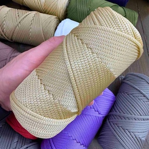 May include: A hand holding a spool of golden-colored yarn. Other spools of yarn in various colors, including purple, green, and gray, are visible in the background. The yarn appears to be tightly wound and ready for crafting projects.
