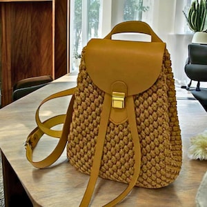 May include: A brown crocheted backpack with a leather flap and buckle closure. The backpack has adjustable straps and a small pocket on the front.