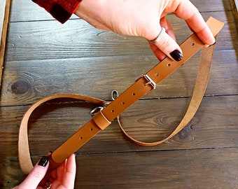 May include: A brown leather camera strap with a silver buckle and a metal ring. The strap is adjustable and has a loop for attaching to a camera.