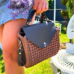 May include: A brown crochet handbag with a dark brown leather flap and handle. The bag features a silver chain and a detachable strap. The bag is held in front of a blurred outdoor background.