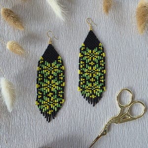 May include: A pair of long, teardrop-shaped beaded earrings featuring a geometric pattern in black, lime green, and yellow seed beads. The earrings hang from gold-toned metal hooks and have a fringe bottom edge. A pair of gold-toned scissors is nearby.