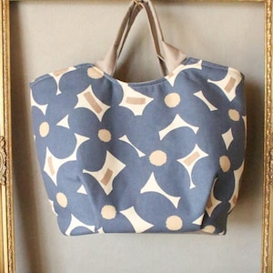 May include: A blue tote bag with a floral pattern of white and tan shapes. The bag has tan handles and is hanging inside a gold ornate picture frame. The background is a neutral gray.