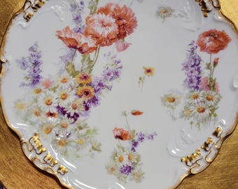 Könnte beinhalten: Ein weißer Porzellanteller mit gewelltem Rand und Goldverzierung. In der Mitte befindet sich ein farbenfrohes Blumenarrangement aus orangefarbenen Mohnblumen, violetten Ritterspornen und weißen Gänseblümchen mit geprägtem Goldrand.