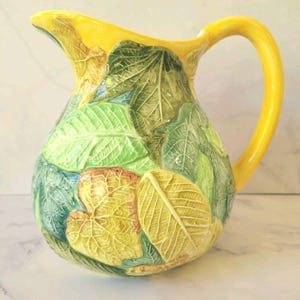 May include: A yellow ceramic pitcher with a leaf design. The pitcher has a rounded body and a curved handle. The leaves are in shades of green, yellow, and brown. The rim and handle are bright yellow.
