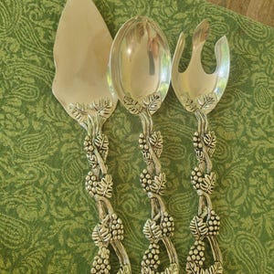 May include: A three-piece silver-toned serving set consisting of a cake server, a large spoon, and a salad fork. Each utensil features a detailed handle design depicting intertwined grapevines with textured leaves and clusters of grapes in relief.