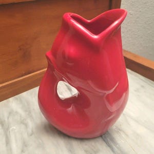 May include: A glossy, bright red ceramic pitcher featuring a distinctively curved, organic silhouette with a wide, heart-shaped pouring spout and a singular, integrated handle opening on the side, showcasing a smooth, reflective surface finish.
