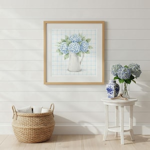 May include: A square framed wall art print featuring a watercolor illustration of blue hydrangea flowers in a white pitcher against a light blue plaid background, displayed alongside a blue and white ceramic vase with fresh blue hydrangeas.