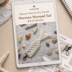 May include: A tablet displays a macrame mermaid tail keychain tutorial. The keychain is white, with a braided body and a fringed tail. Seashells and starfish surround the tablet. The text on the tablet reads "Beginner Step-by-Step Tutorial Macrame Mermaid Tail KEYCHAIN."