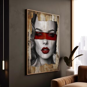 Op de afbeelding: Een zwart-wit portret van een vrouw met rode lippen en een rode band die haar ogen bedekt. Het portret is ingelijst in een houten lijst met een verweerde look.