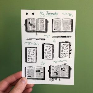 May include: A white card with black ink illustrations of A5 journals. The card has the text "A5 Journals by @georgiajournals" at the top. The illustrations include open journals with pages, pens, and stars.