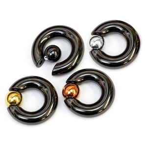 May include: Four circular captive bead rings with a polished black finish. Each ring features a single removable ball bead in the center. The beads are finished in metallic black, silver, gold, and copper tones, creating a distinct color contrast.