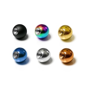 Puede incluir: Seis esferas metálicas en varios colores: negro, iridiscente, dorado, azul, plateado y cobrizo. Cada esfera tiene una hendidura circular. Las esferas están dispuestas sobre un fondo blanco.