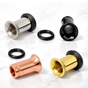 May include: An assortment of metal ear gauges in silver, black, copper, and gold. Each gauge has a flared end and a black rubber O-ring. The gauges are displayed on a white marble surface.