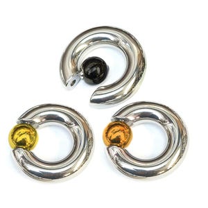 Könnte beinhalten: Drei silberfarbene, kreisförmige Körperschmuckstücke mit poliertem Finish. Jedes Stück hat einen kugelförmigen Akzent in Schwarz, Gold und Orange. Der Schmuck ist mit einer Scharnieröffnung zum Einsetzen versehen.