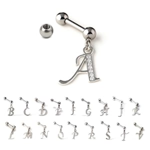 May include: A collection of silver-colored barbell-style body jewelry with dangling initial charms. The letter "A" is shown in detail, with a clear gem-studded design. Additional charms with letters B through V are also displayed.