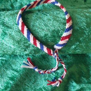 Könnte beinhalten: Ein rot-weiß-blaues Freundschaftsarmband mit einem geflochtenen Knoten am Ende.