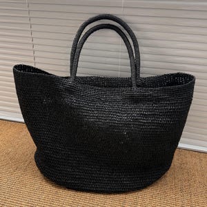 Pode incluir: Uma grande sacola preta tecida com duas al&ccedil;as arredondadas. A sacola tem uma parte superior larga e aberta e uma base arredondada. O material tecido tem uma apar&ecirc;ncia texturizada. A sacola est&aacute; sobre uma superf&iacute;cie marrom.