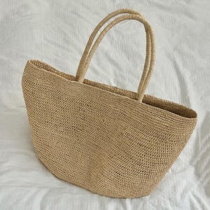 Pode incluir: Uma caneca de cer&acirc;mica branca com uma al&ccedil;a preta e o texto "But First, Coffee" em negrito preto. A caneca est&aacute; sobre um fundo desfocado, sugerindo um ambiente acolhedor.