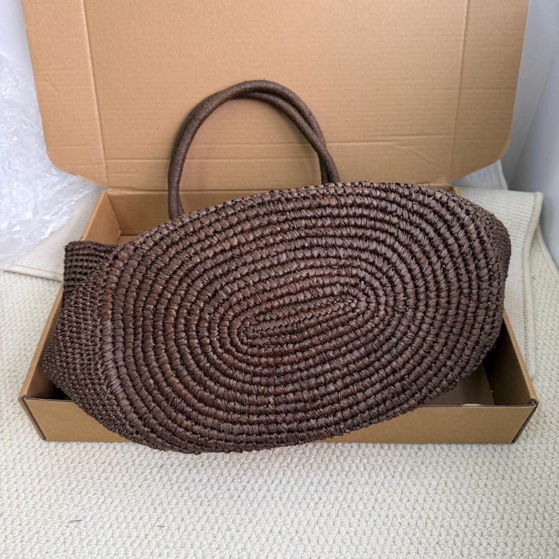 Pode incluir: Uma sacola tote marrom tecida com duas al&ccedil;as, dentro de uma caixa de papel&atilde;o. A sacola tem uma base oval e um padr&atilde;o de tecido em espiral. As al&ccedil;as tamb&eacute;m s&atilde;o tecidas e parecem resistentes. A sacola &eacute; de um tom natural e terroso.