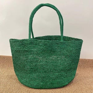 May include: A green woven tote bag with two matching handles. The bag is made of a textured material and has a rounded shape. The bag is empty and sits on a neutral-colored surface.