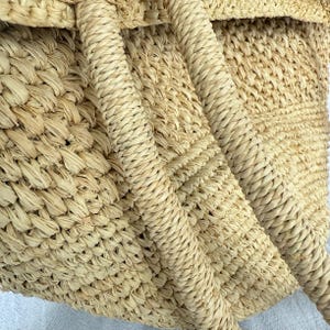 May include: Close-up of a woven straw tote bag. The bag has a natural beige colour with a textured weave. The handles are made of tightly wound straw, adding to the bag's rustic aesthetic. The bag's design suggests it is suitable for carrying items.