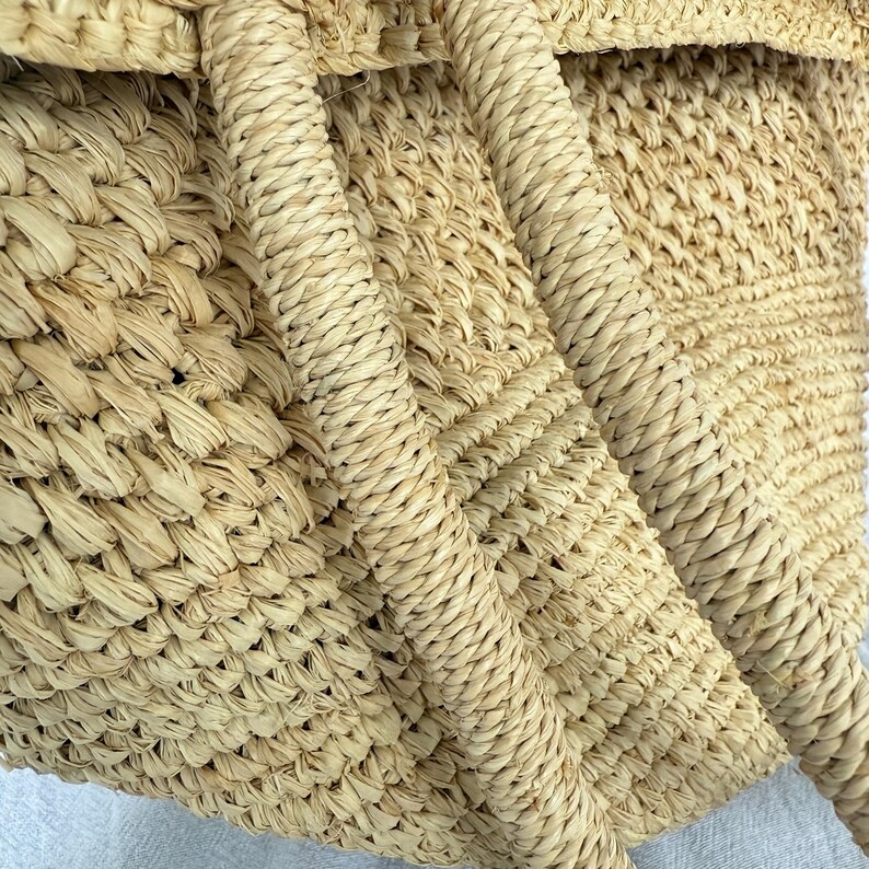 May include: Close-up of a woven straw tote bag. The bag has a natural beige colour with a textured weave. The handles are made of tightly wound straw, adding to the bag's rustic aesthetic. The bag's design suggests it is suitable for carrying items.