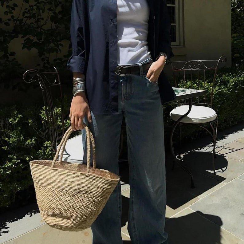 May include: A woven tote bag in light brown is held. The person is wearing a navy blue shirt over a white top and light blue jeans. Silver bracelets and a black belt are also visible. The setting is outdoors, with a wrought iron chair and table in the background.