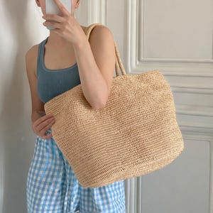 Pode incluir: Uma sacola tote de palha tecida com al&ccedil;as resistentes. A sacola &eacute; de cor bege claro e tem formato retangular. A sacola est&aacute; sendo segurada por uma pessoa vestindo uma regata azul e cal&ccedil;as xadrez azuis e brancas.