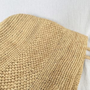 May include: A beige woven straw tote bag. The bag has a rectangular shape with a textured surface and two handles. The weave pattern is visible, creating a detailed design. The bag is made of natural materials.