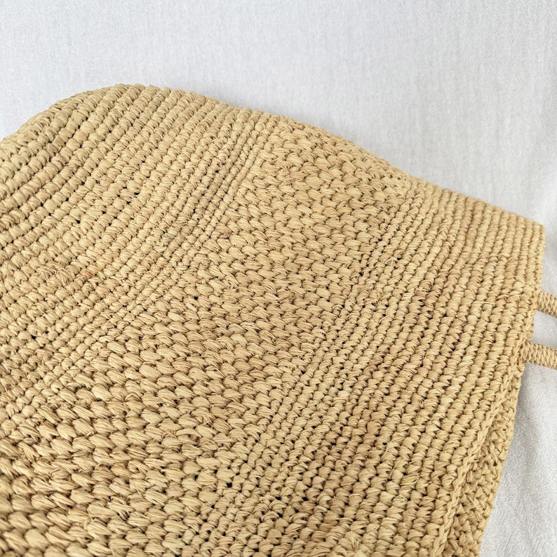 May include: A beige woven straw tote bag. The bag has a rectangular shape with a textured surface and two handles. The weave pattern is visible, creating a detailed design. The bag is made of natural materials.