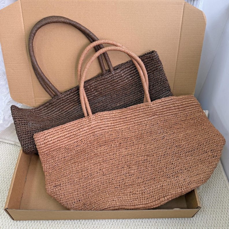 Pode incluir: Duas sacolas tote tecidas em tons de castanho. Uma &eacute; marrom chocolate mais escuro, e a outra &eacute; de cor castanha mais clara. Ambas as sacolas t&ecirc;m al&ccedil;as tecidas combinando e s&atilde;o de formato retangular. Elas est&atilde;o dentro de uma caixa de papel&atilde;o.