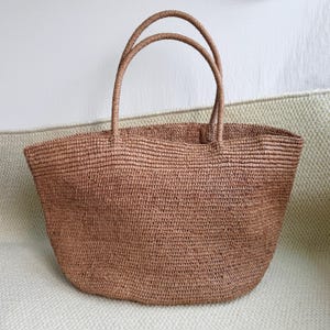 Pode incluir: Uma sacola tote marrom tecida com duas al&ccedil;as arredondadas. A sacola &eacute; feita de um material de fibra natural e tem uma parte superior larga e aberta. A sacola est&aacute; em uma superf&iacute;cie verde clara.